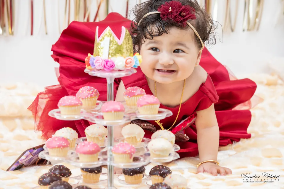 A smiling baby dressed in red beside a cake and cupcakes